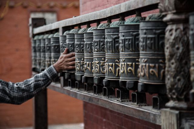 Image of a person running their hand along a line of religious prayer wheels in a temple. Gold engravings on the wheels are mantras used for spiritual and meditative practices in Buddhism. Ideal for content related to spirituality, culture, religious traditions, meditation practices, faith, travel destinations involving temples and cultural sites.