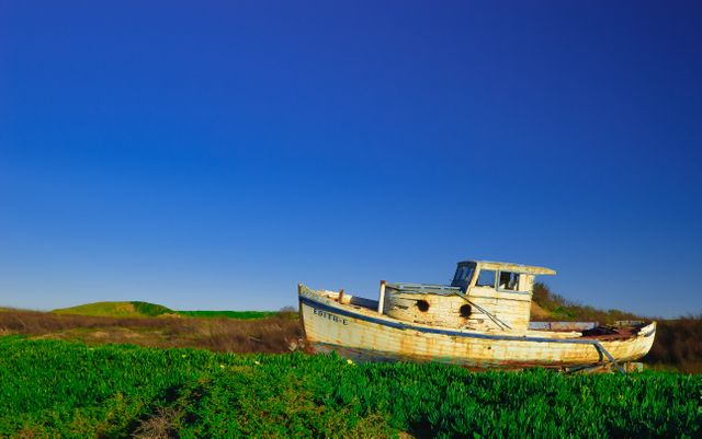Depicts an abandoned, rusty boat sitting in an overgrown field under a clear, blue sky. Useful for themes of abandonment, decay, nature reclaiming human artifacts, and maritime history. Suitable for environmental articles, background visuals in films and documentaries, and artistic reference for decay and nature themes.