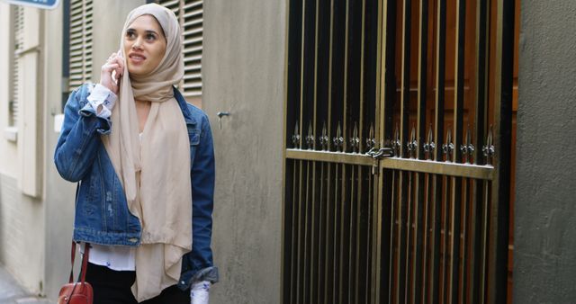 Young Muslim woman wearing hijab talking on mobile phone while walking along urban street. Ideal for use in articles about urban life, communication, diversity, fashion, and culture. Possible use in advertisements for mobile phones or fashion industry targeted at younger demographics.