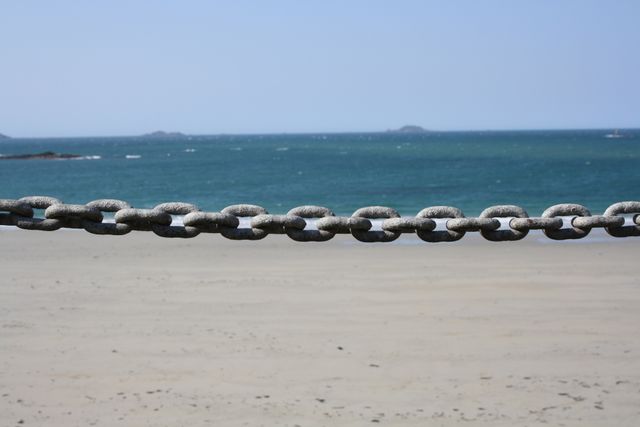 This image highlights a close-up view of a weathered, rusty chain stretched in the foreground, framing a serene sandy beach with calm ocean waters extending to the horizon. Use for themes such as coastal scenery, nautical elements, tranquility, or summer. Ideal for websites, travel brochures, and environmental articles focusing on the beauty and serenity of coastal landscapes.
