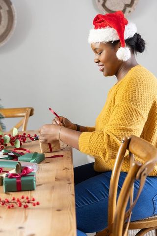 African american woman wrapping christmas gifts at dining table wearing santa hat