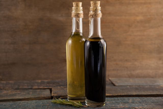 This image shows two glass bottles, one containing olive oil and the other balsamic vinegar, placed on a rustic wooden table. The bottles have cork stoppers and are tied with twine, adding a touch of rustic charm. Ideal for use in culinary blogs, cooking websites, healthy eating articles, and organic food promotions.
