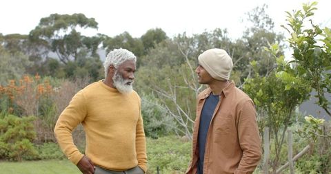 Mature Black and South Asian men chatting in garden wearing mustard sweater and beanie