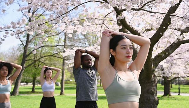 Asian woman stretching under cherry blossom trees with diverse group doing outdoor fitness