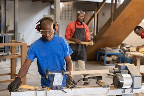 African american carpenter carving wooden spindle on lathe in busy woodworking workshop