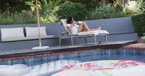 African american woman relaxing on poolside chaise reading with sunglasses, summer leisure