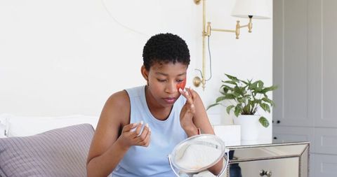 African american woman applying under-eye skincare on bed in bright minimalist bedroom