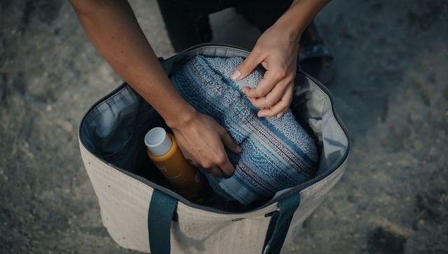Packing beach tote with striped towel and sunscreen on pebbled shore overhead