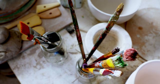Assorted paintbrushes standing upright in glass jars on an artistic workspace desk with various painting tools and supplies. Suitable for illustrating creative artistic environments, fostering creativity, DIY projects, art supply store promotions, and blogs or articles about art and painting.
