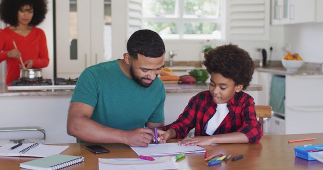 Father assists son with homework at kitchen table, fostering educational growth and family bonding. Meanwhile, mother prepares meal in background, creating warm, supportive home environment. Ideal for depicting family life, education at home, parental involvement, and domestic tranquility.