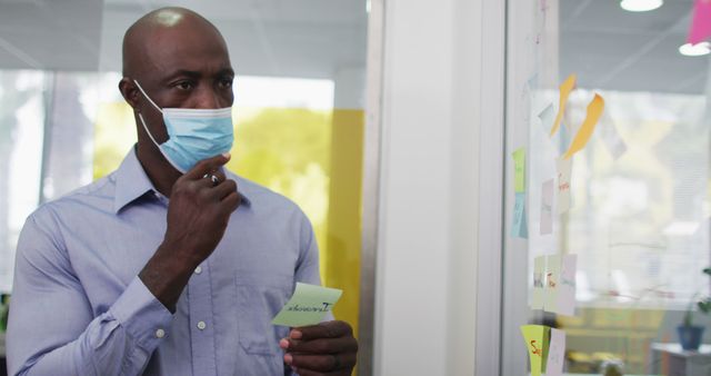 Man analyzing ideas and plans on sticky notes attached to a glass board in an office. He is wearing a face mask. Perfect for illustrating work during a pandemic, office planning sessions, brainstorming techniques, and business strategy discussions.
