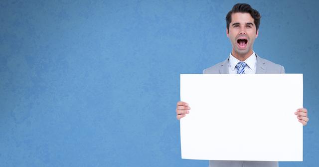 Young businessman in a suit screaming while holding an empty billboard with a vivid blue background. Ideal for use in corporate presentations, advertising, announcements, or promotional materials requiring a dramatic and engaging expression.
