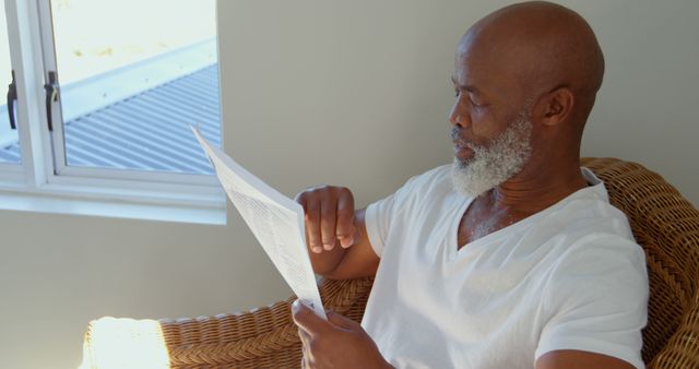Side view of black mature man reading newspaper at home. He is relaxing on armchair 4k