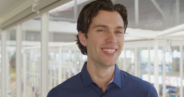 Young man smiling confidently in modern office setting wearing a blue shirt. This image is suitable for corporate communication, professional profiles, employee spotlight features, and modern business presentations.