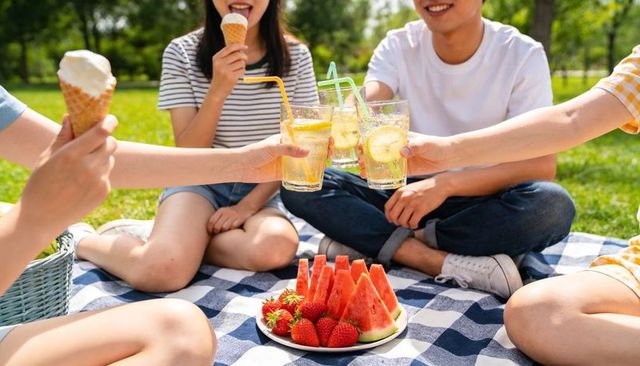 Friends toasting lemonade at summer picnic with watermelon, strawberries and ice cream