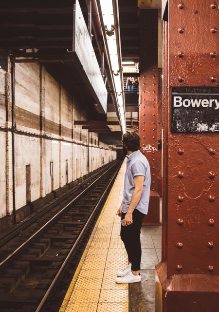 Individual stands on subway platform, gazing down tracks at the Bowery Station. Utilizable for themes like urban life, travel, public transportation, commuting, city living, and exploration.