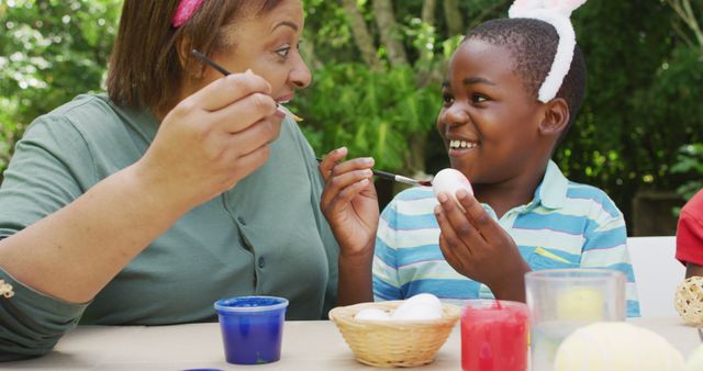 Smiling mother and son painting Easter eggs at outdoor table, enjoying quality time together. Green foliage in background suggesting holiday season and springtime environment. Suitable for depicting family activities, Easter celebrations, and bonding moments.