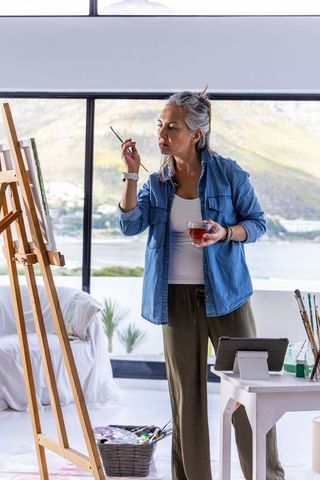 Senior asian woman painting at easel in bright waterfront studio holding brush and tea
