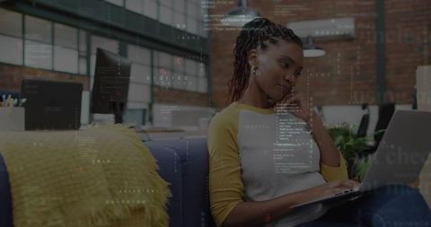Woman coding on laptop while leaning on couch in sunlit modern open-plan coworking office