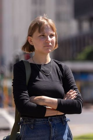 Young woman standing arms crossed on sunlit urban street wearing black top and denim backpack strap