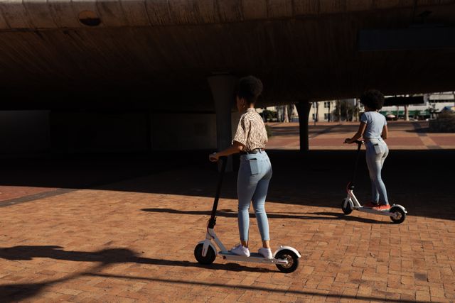 Side view of two biracial twin sisters enjoying free time together in a city on a sunny day, riding electric scooters in an urban park.