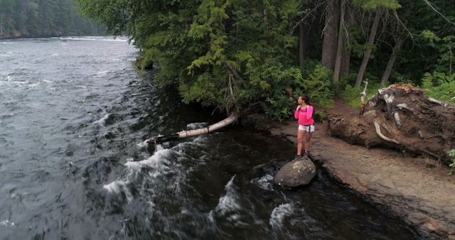 Young woman standing on rock along riverbank surrounded by dense forest, wearing summer hiking gear and gazing at the rushing water. This image can be used for promoting outdoor adventures, travel blogs, hiking tours, nature preservation campaigns, summer vacation planning, and active lifestyle events.