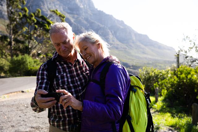 Senior couple enjoying a hike in a mountainous area, taking a break to use a smartphone. Ideal for promoting active lifestyles, travel, adventure, and technology use among older adults. Perfect for retirement planning, outdoor activities, and healthy living campaigns.