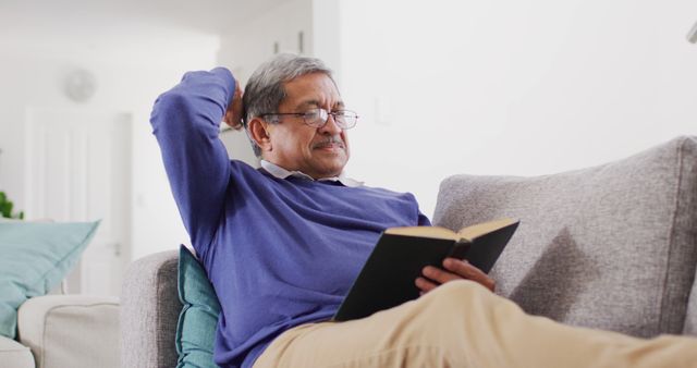 Senior man enjoying book while sitting on comfortable sofa in home. Suitable for promoting retirement living, leisure activities, literature, peaceful home environments, senior lifestyle.