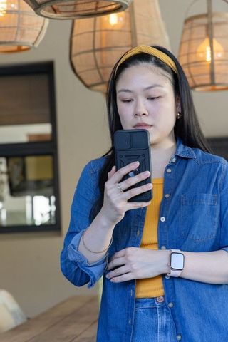 Asian woman standing in cozy cafe holding phone wearing denim shirt and smartwatch