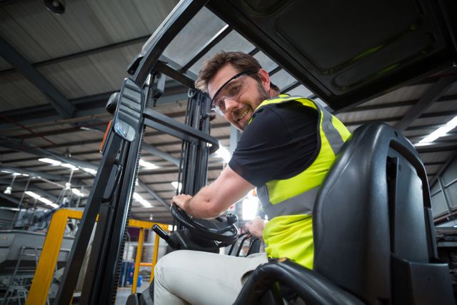 This image depicts a male factory worker smiling while driving a forklift in an industrial warehouse. He is wearing a safety vest and goggles, emphasizing the importance of occupational safety. This image can be used for promoting workplace safety, illustrating industrial operations, or highlighting logistics and transportation in manufacturing environments.