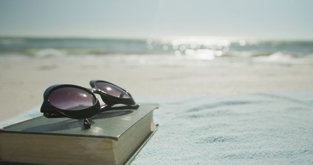 Sunglasses are placed on a closed book on a beach towel with the ocean in the background. The scene evokes relaxation and leisure, typical of a beach vacation. Perfect for travel blogs, advertisements for vacation packages, or promotions related to summer or beach activities.