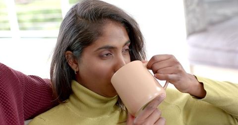 Relaxing Indian woman sipping from beige mug on couch wearing green turtleneck sweater