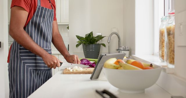 Man slicing red cabbage and onions in modern kitchen, referencing recipe on tablet. Useful for cooking blogs, dietary guides, technological kitchen gadgets, smart kitchen setups, healthy living tips and home cooking tutorials.