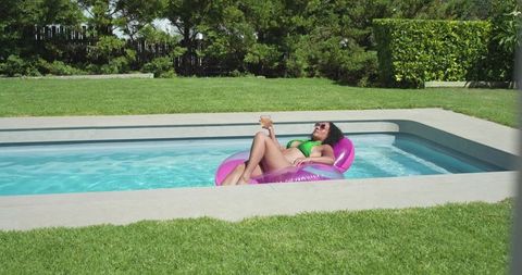 Sunlit african woman relaxing on pink inflatable ring in backyard pool with drink