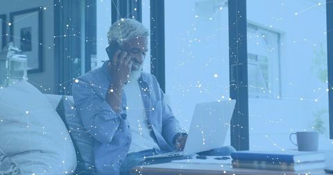 Senior man working remotely on laptop while talking on smartphone at home office