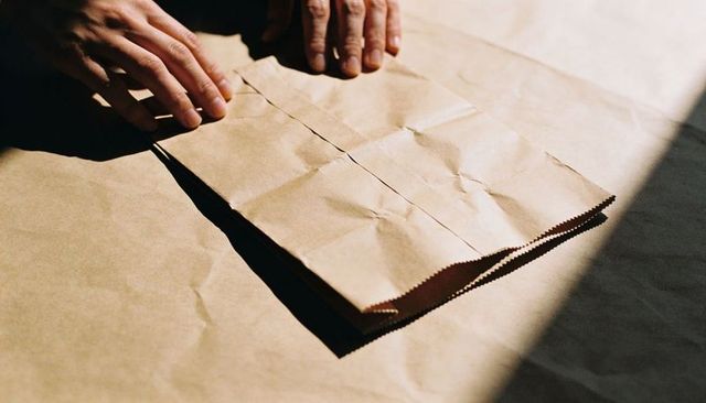 Hands smoothing folded kraft paper bag on sunlit tabletop with tactile texture and shadow