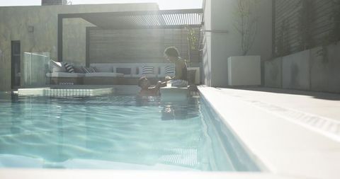 Sunlit couple relaxing at modern pool holding hands near pergola and glass barrier