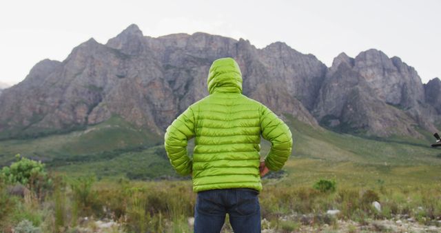 Individual wearing bright green jacket standing in rugged outdoor setting, facing tall, rocky mountains. Ideal for use in adventure blogs, travel advertisements, outdoor gear promotions, and environmental awareness campaigns. Evokes themes of exploration, solitude, and appreciation for natural landscapes.