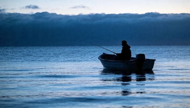 Solitary angler fishing at dawn in small boat silhouette on calm foggy sea