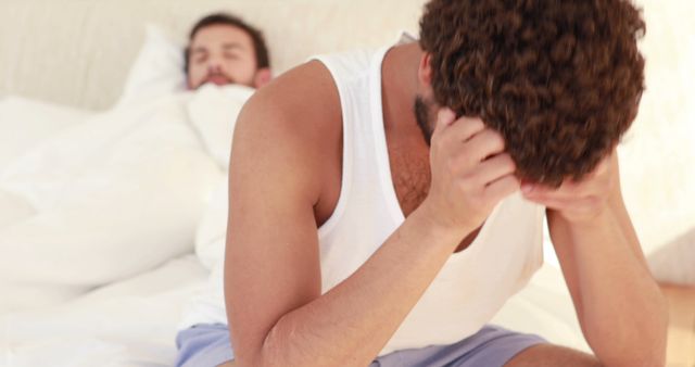 Man holding his head in frustration while sitting on bed beside his sleeping partner, indicating trouble or conflict in relationship. Useful for depicting relationship issues, emotional stress, communication problems, or mental health scenarios in personal life settings.