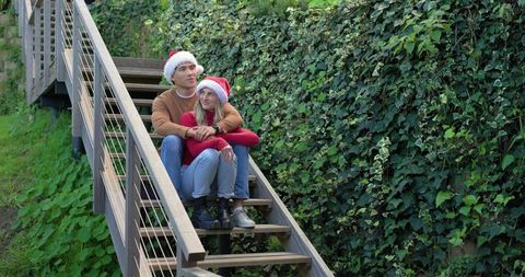 Couple sitting on wooden outdoor stairs wearing santa hats and embracing in garden