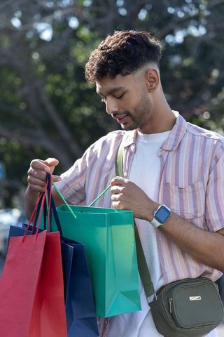 Young indian man inspecting colorful shopping bags in shaded park wearing casual outfit