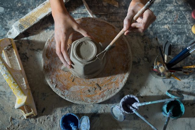 Hands of a female potter carving a clay mug in a pottery workshop. Ideal for use in articles or advertisements about pottery, craftsmanship, creative hobbies, or artisan work. Can also be used in educational materials about traditional crafts and ceramic art.
