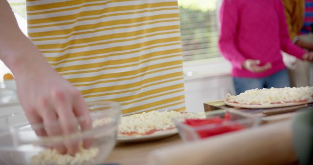 Person adding cheese to pizzas in a kitchen with striped shirt in foreground. Family cooking activity is visible in the background. Useful for cooking tutorials, family activities, or food blogs focusing on homemade recipes.