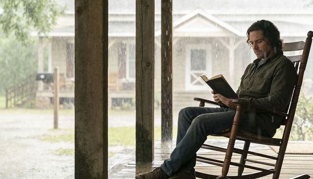 Man reading on rustic porch rocker during heavy rainstorm, rural solitude and cozy shelter