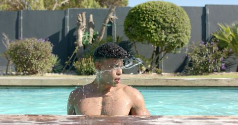 African american man rising from backyard pool, splashing water over face, sunny swim