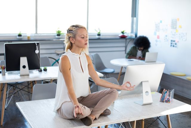 Female executive practicing meditation on desk in a modern office. Ideal for illustrating workplace wellness, stress relief, and mindfulness in a corporate setting. Useful for articles on mental health, productivity, and creating a balanced work environment.