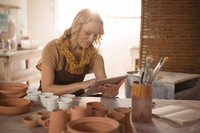Female potter in bright workshop using digital tablet, surrounded by clay pots and artisan tools. Ideal for concepts of art and technology, modern craftsmanship, small business productivity, and creative workspaces.