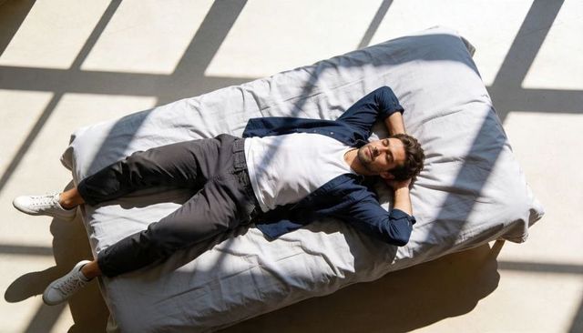 Young man resting on minimalist mattress under sunlit window grid, overhead lifestyle composition