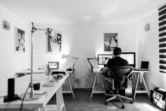 Man working on a computer in a clean, modern home office with two monitors, surrounded by contemporary decor and office equipment. Ideal for illustrating home office setups, remote work environments, productivity concepts, and modern workspace designs.